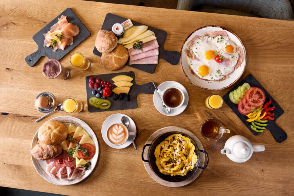 Ein reichhaltiges Frühstücksarrangement mit verschiedenen Brötchen, Käse, Schinken, frischem Obst, Eiern und Kaffee auf einem Holztisch.