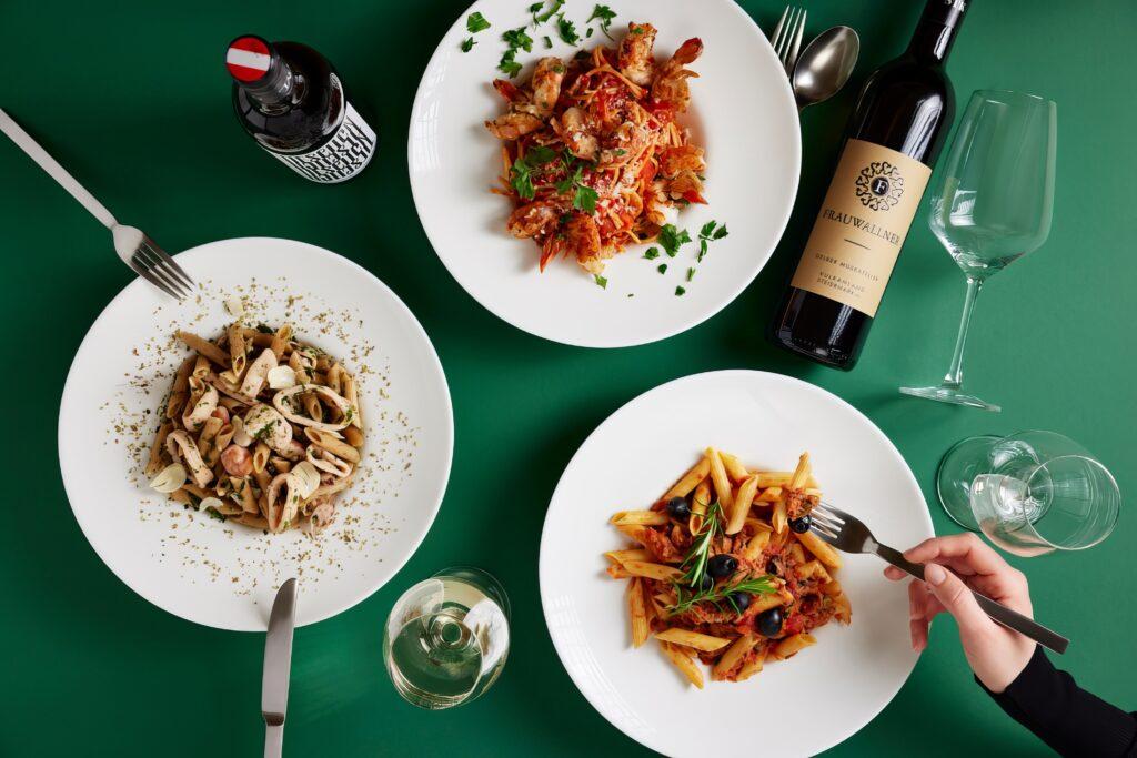 Verschiedene Pasta-Gerichte auf einem grünen Tisch mit Wein und Gläsern.